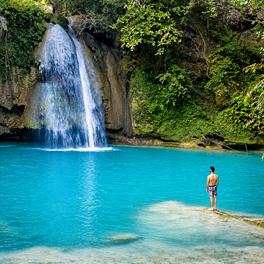 A visitor standing at the turquoise pool of Kawasan Falls